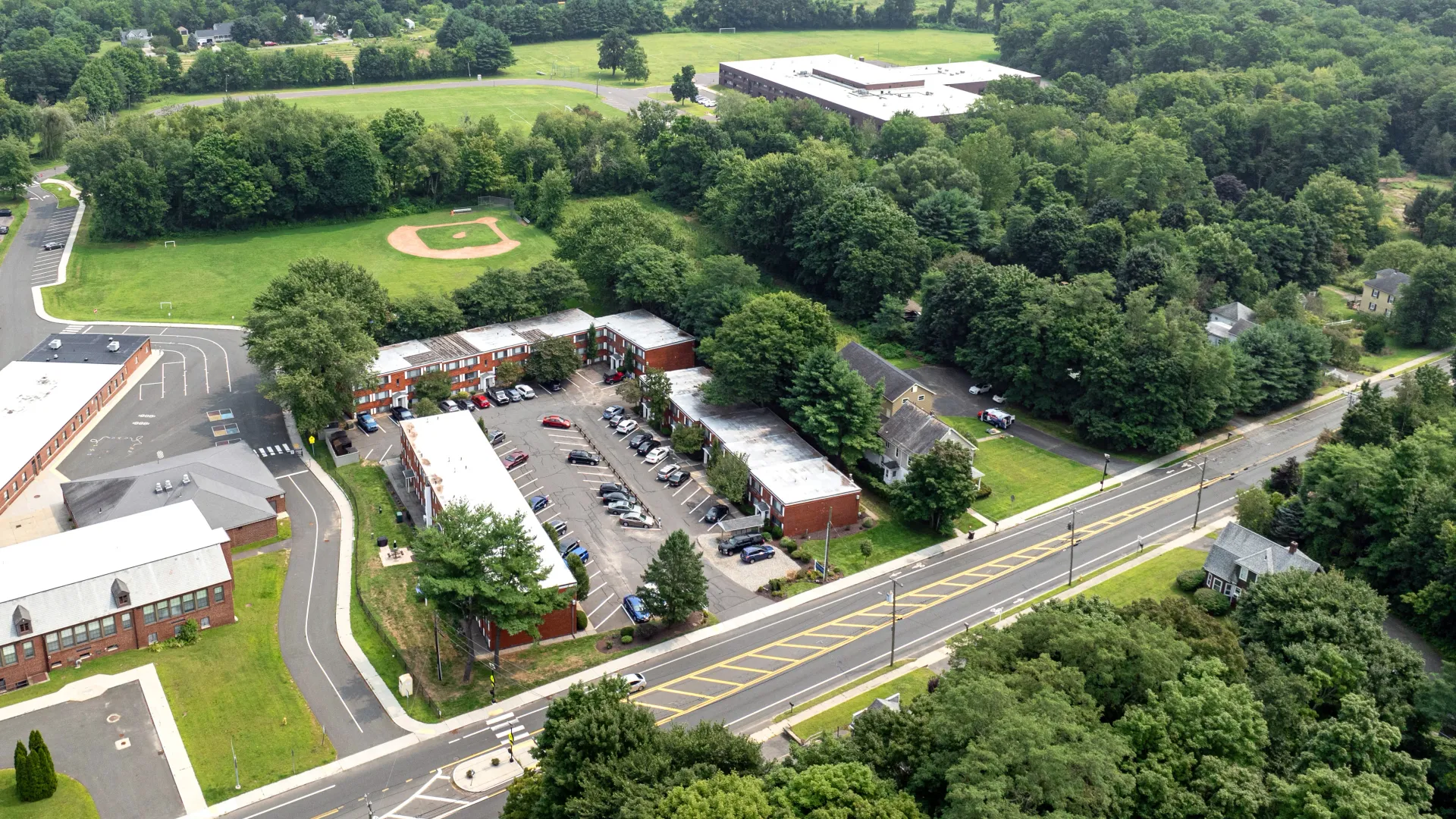 Aerial view of Country Manor Apartments, featuring multiple apartment buildings, parking lots, green spaces, and a nearby baseball field, all surrounded by lush trees and residential areas.