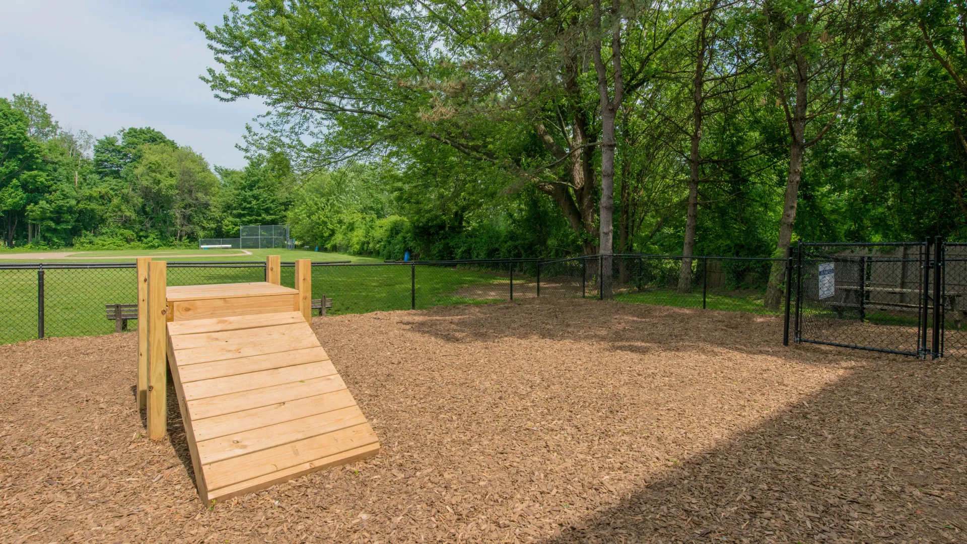 Fenced dog park at Country Manor Apartment Homes with agility equipment, wood-chip ground cover, and surrounding green space and trees.