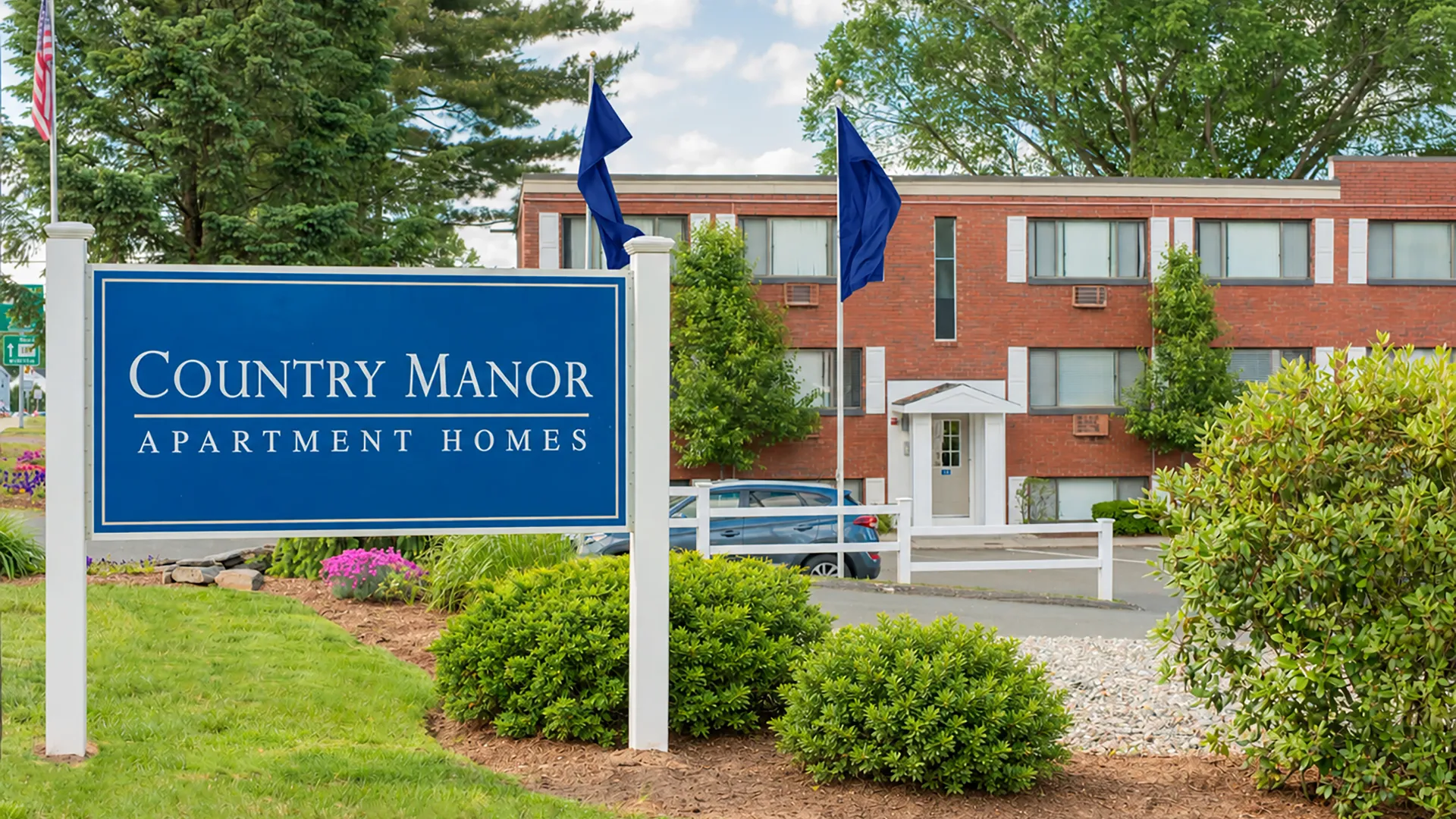 Blue Country Manor Apartment Homes sign surrounded by landscaped gardens and greenery at the community entrance.