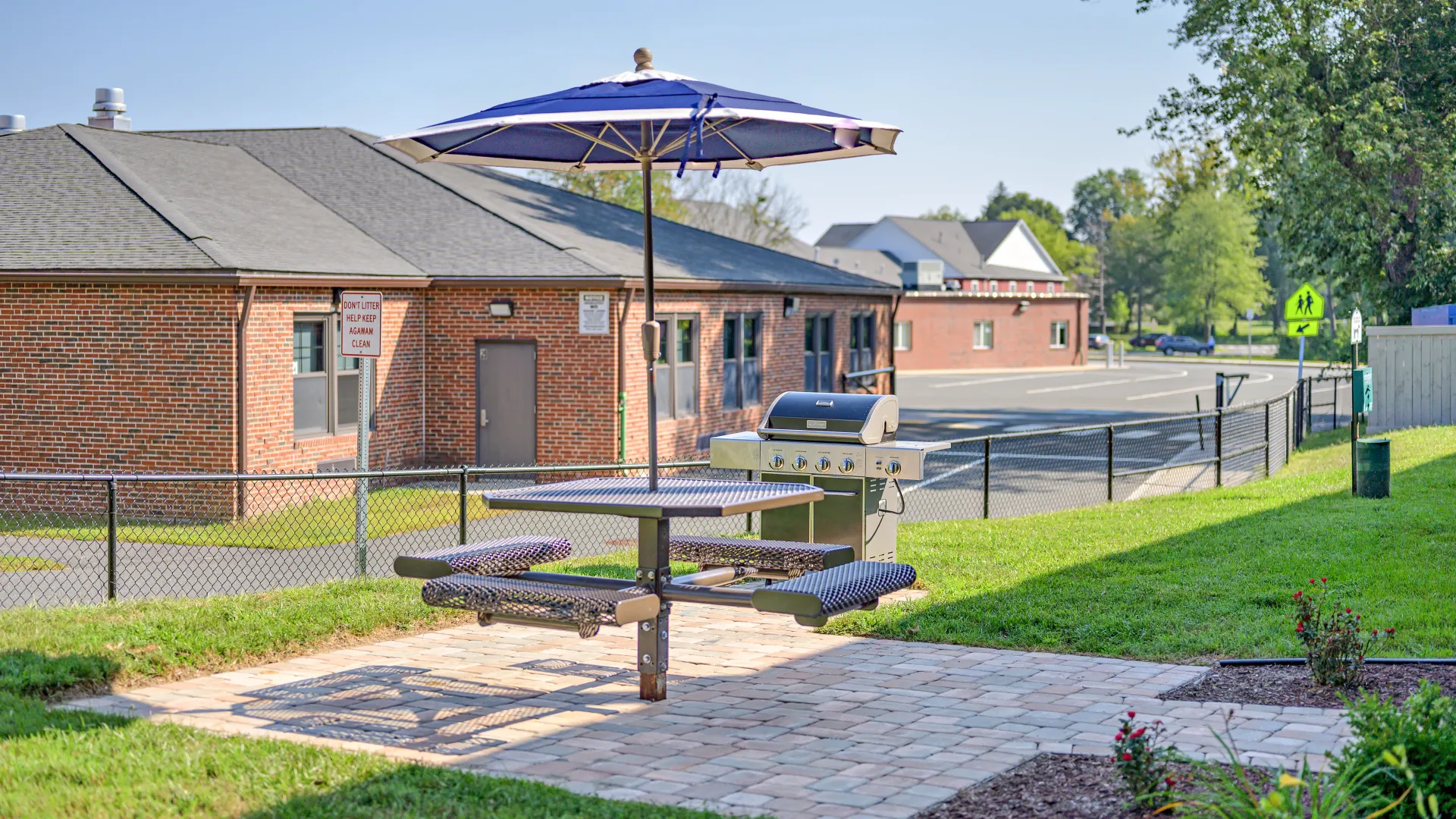 Outdoor picnic area at Country Manor Apartment Homes with a shaded picnic table, gas grill, and paved patio surrounded by green lawn.