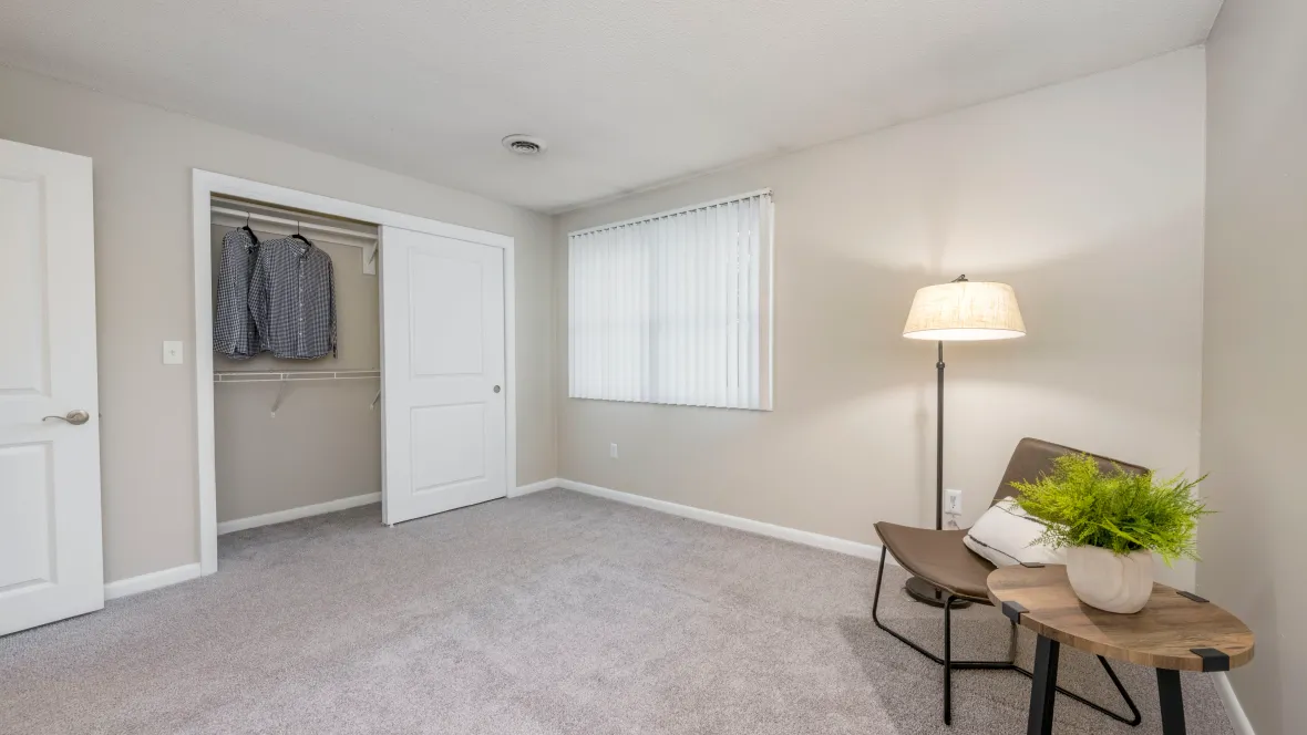 A roomy bedroom with plush carpeting, a large window with privacy blinds, and a generous closet equipped with built-in organizers.