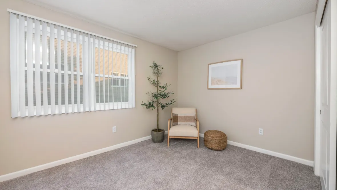 A large bedroom with soft, cozy carpeting, bathed in natural light from a large window, complete with privacy blinds.