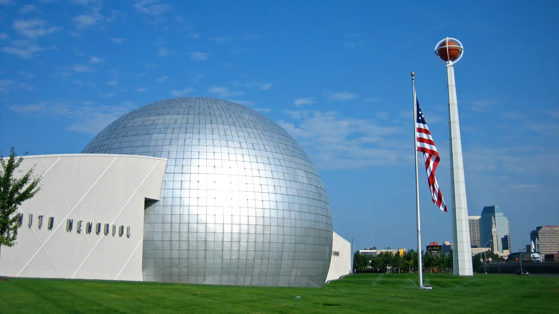 A photo of the Basketball Hall of Fame, featuring a large silver dome, an American flag, and a tall tower with a basketball on top against a clear blue sky.