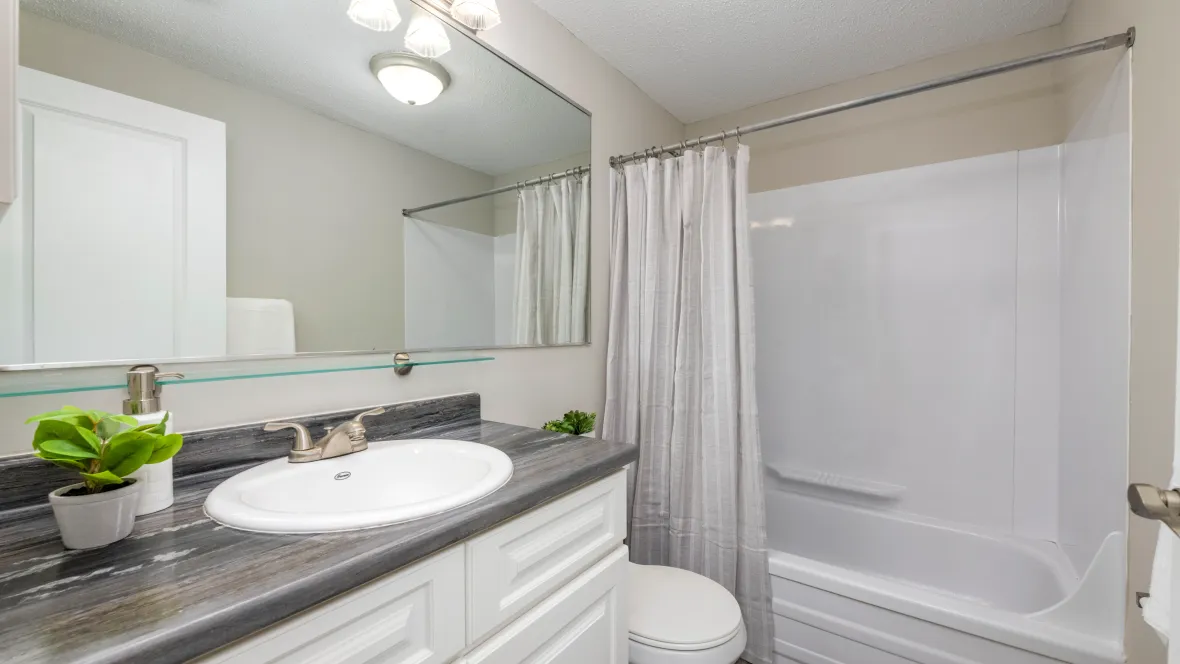 Charming bathroom with refined elements, including an expansive mirror, white cabinetry, bright lighting, a shower/tub combo, and black fusion countertops.