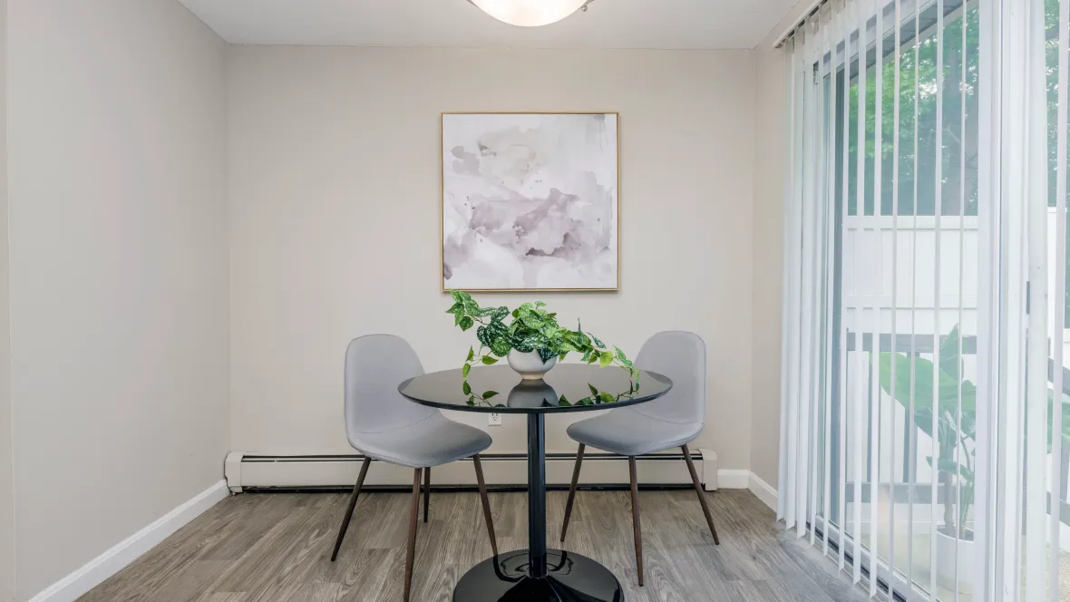 Stylish dining area with a table and chairs next to sliding patio doors with privacy blinds. An overhead light fixture and cozy baseboard floor heater enhance comfort.