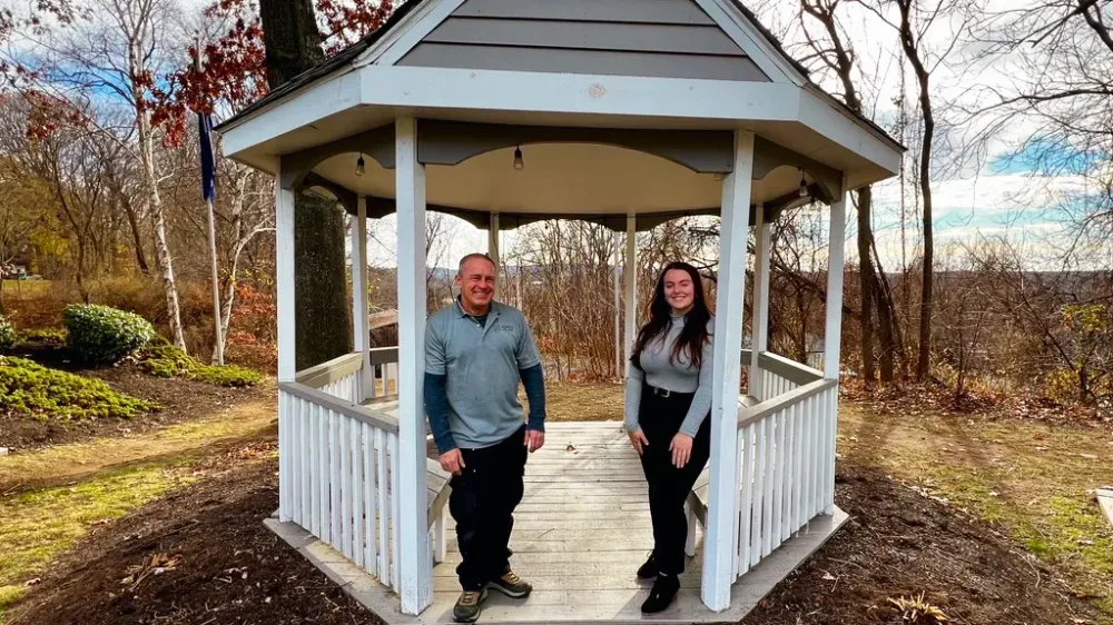 Two team members standing under the white gazebo at The Willows Apartments on a fall day with trees and landscaping in the background.