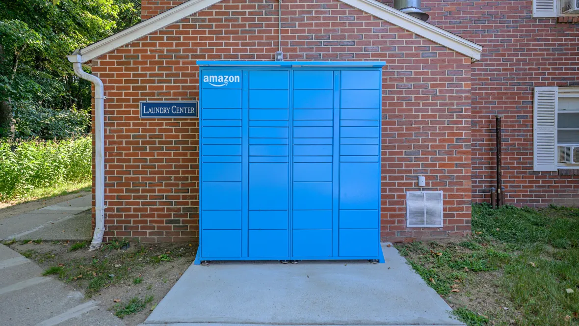 A vibrant blue Amazon Hub package locker outside in front of a red brick apartment building ensuring hassle-free, 24/7 package delivery and pickup for residents.