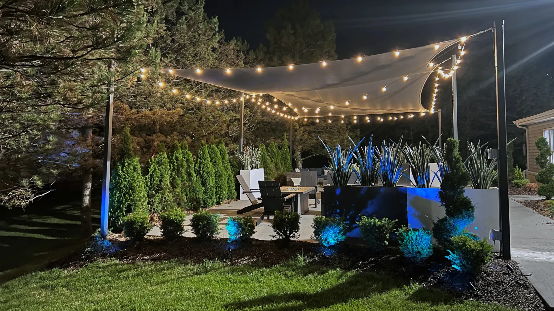 Outdoor lounge at night, bistro lighting and blue landscape uplighting create a serene ambiance for relaxation under the night sky.