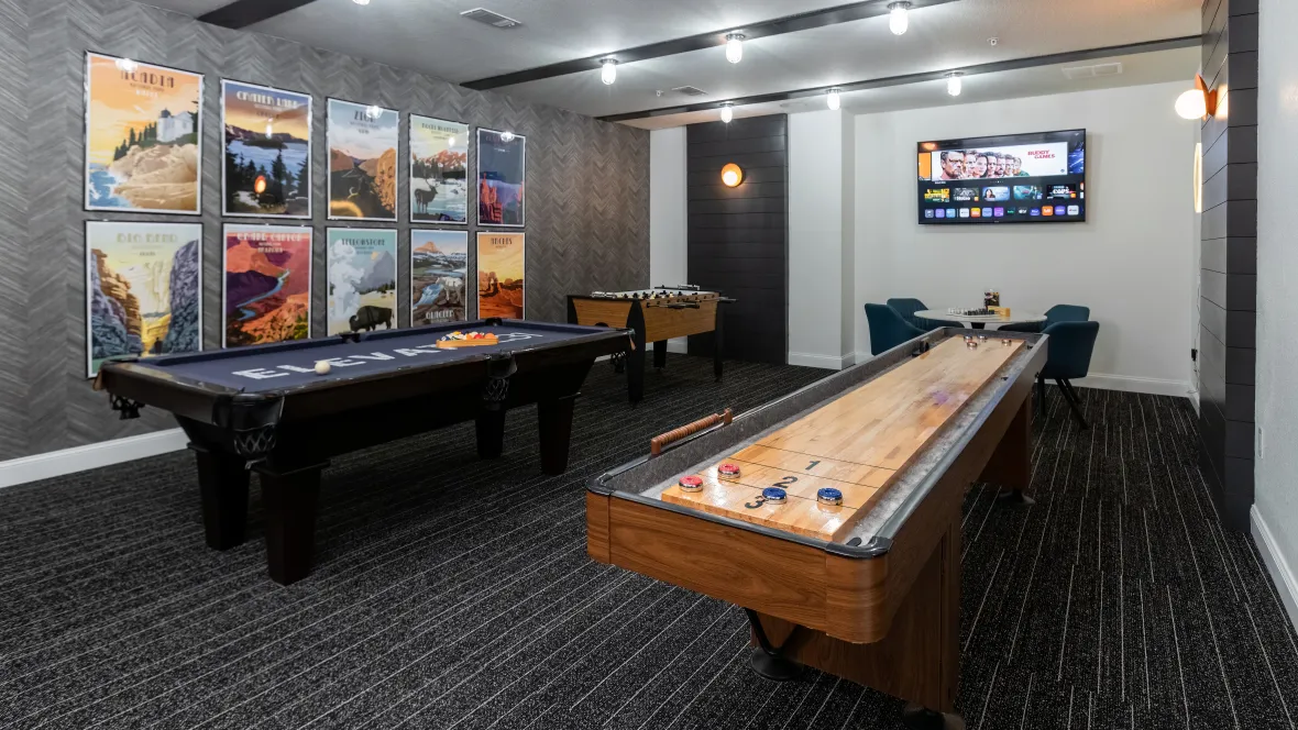 A game room with a small table and four chairs in the corner, with a television mounted on the wall above. The room also features a pool table, shuffleboard table, and foosball table.