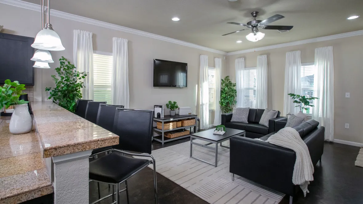 A view of the large living room from the open kitchen space with breakfast bar, showing the full furniture package including sofa, love seat, coffee table, 55-inch television, and ceiling fan.