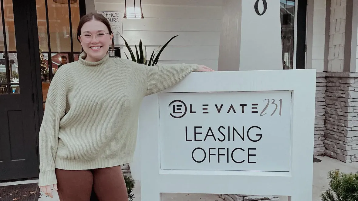 An employee in a tan sweater with their arm on top of a sign that reads "Elevate 231 Leasing Office".