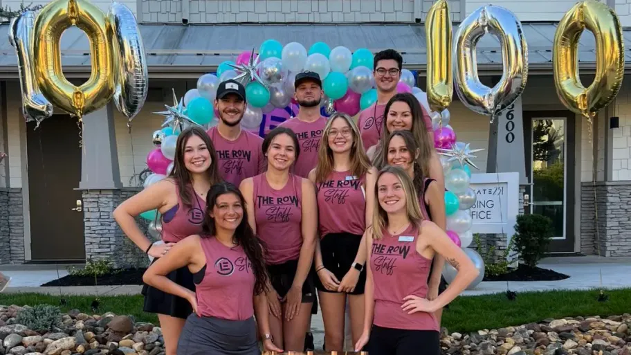 The Row team gathered for a team photo in front of a colorful balloon arc with balloons on either side reading "100".