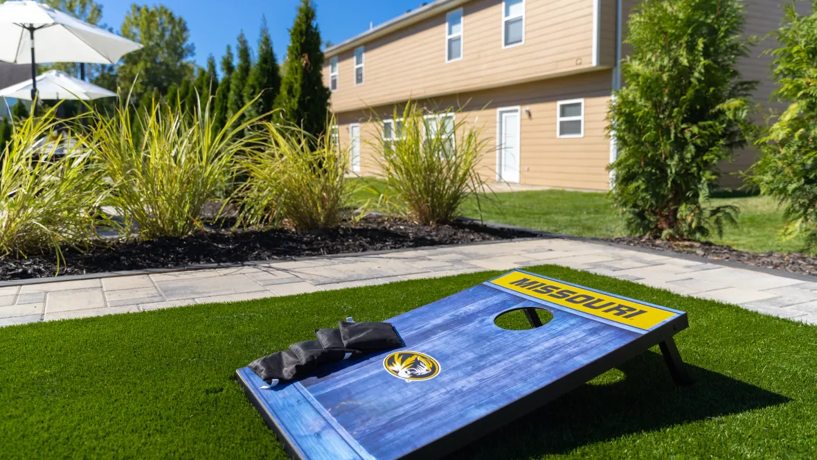 Close-up of a Mizzou-themed cornhole board in the lush courtyard, featuring the University of Missouri's oval tiger head logo.