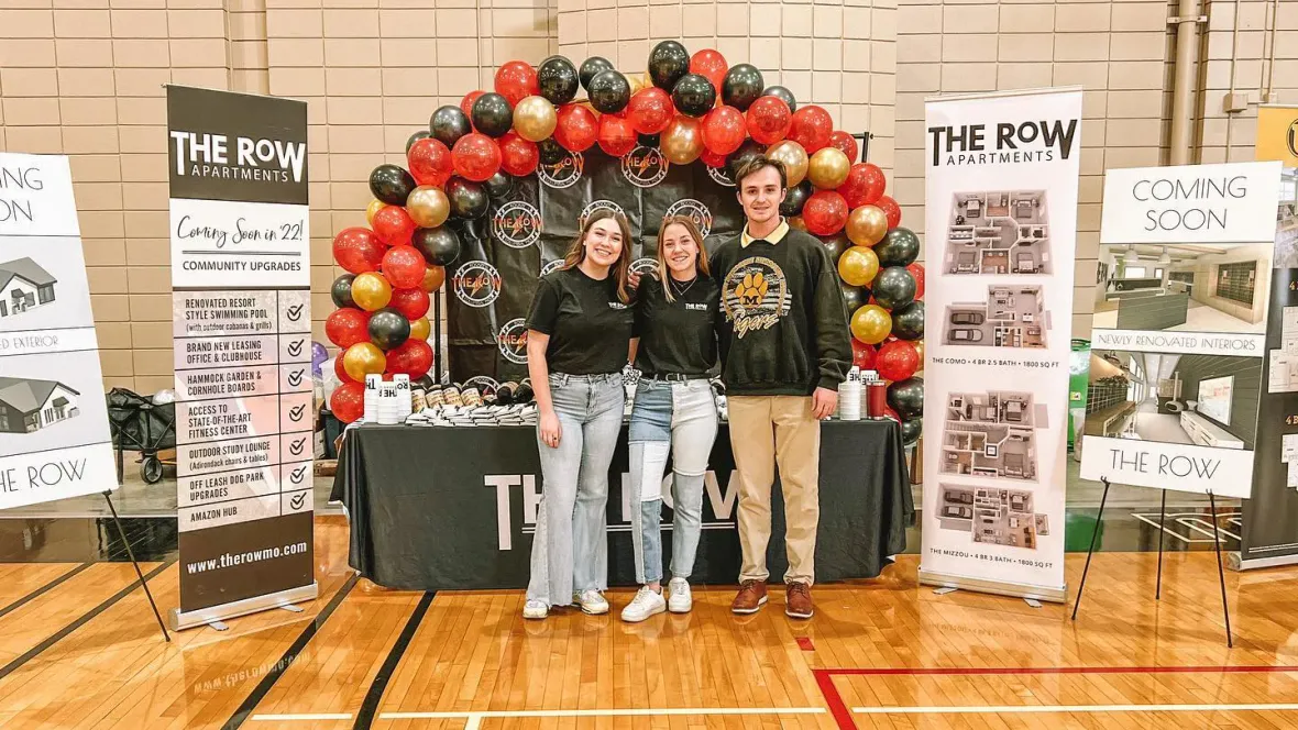 The Row team members standing in front of their housing fair set up in front of a red, gold and black balloon arc.