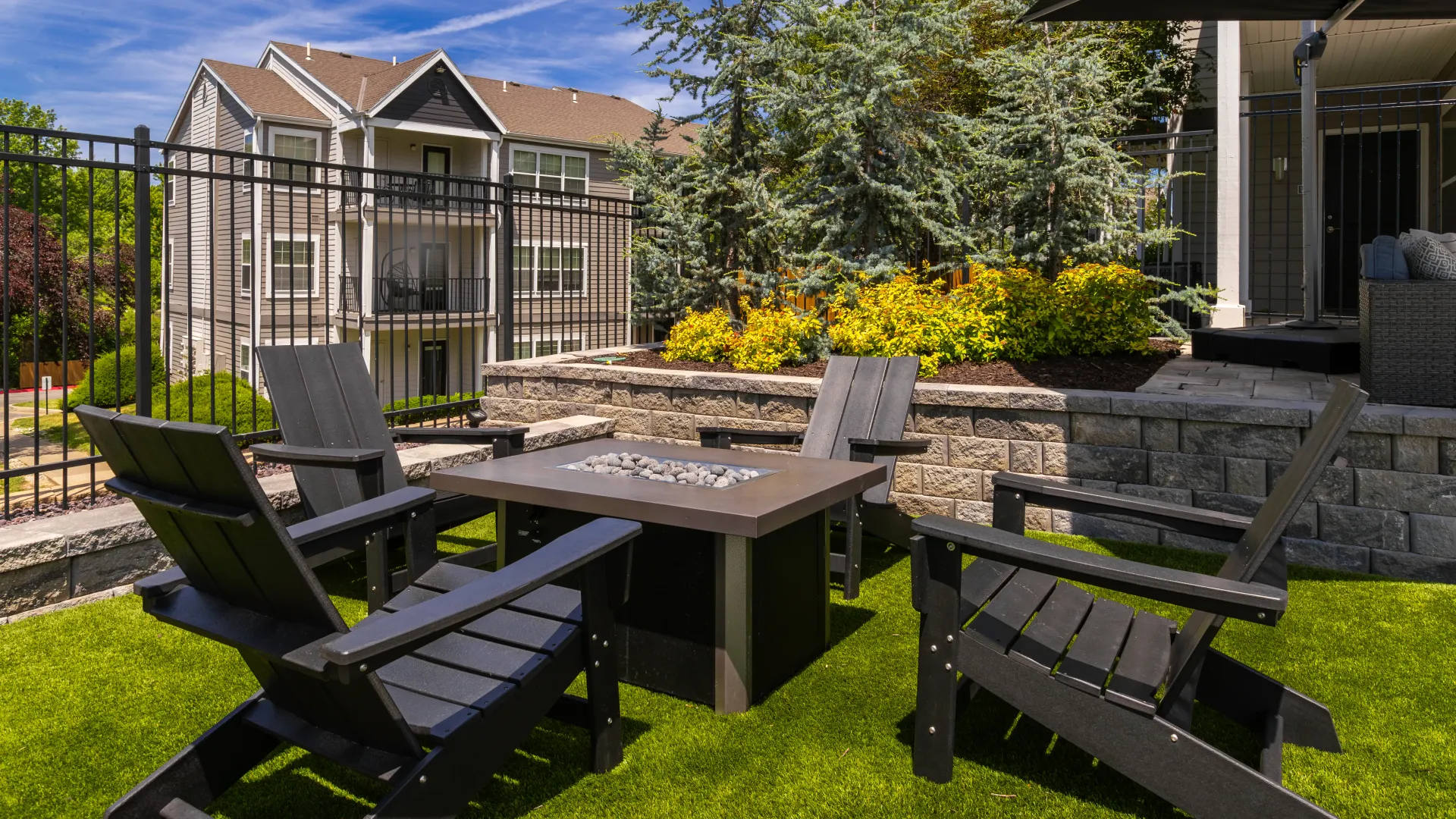 Outdoor fire pit area with black Adirondack chairs, surrounded by lush greenery and adjacent to an apartment building.