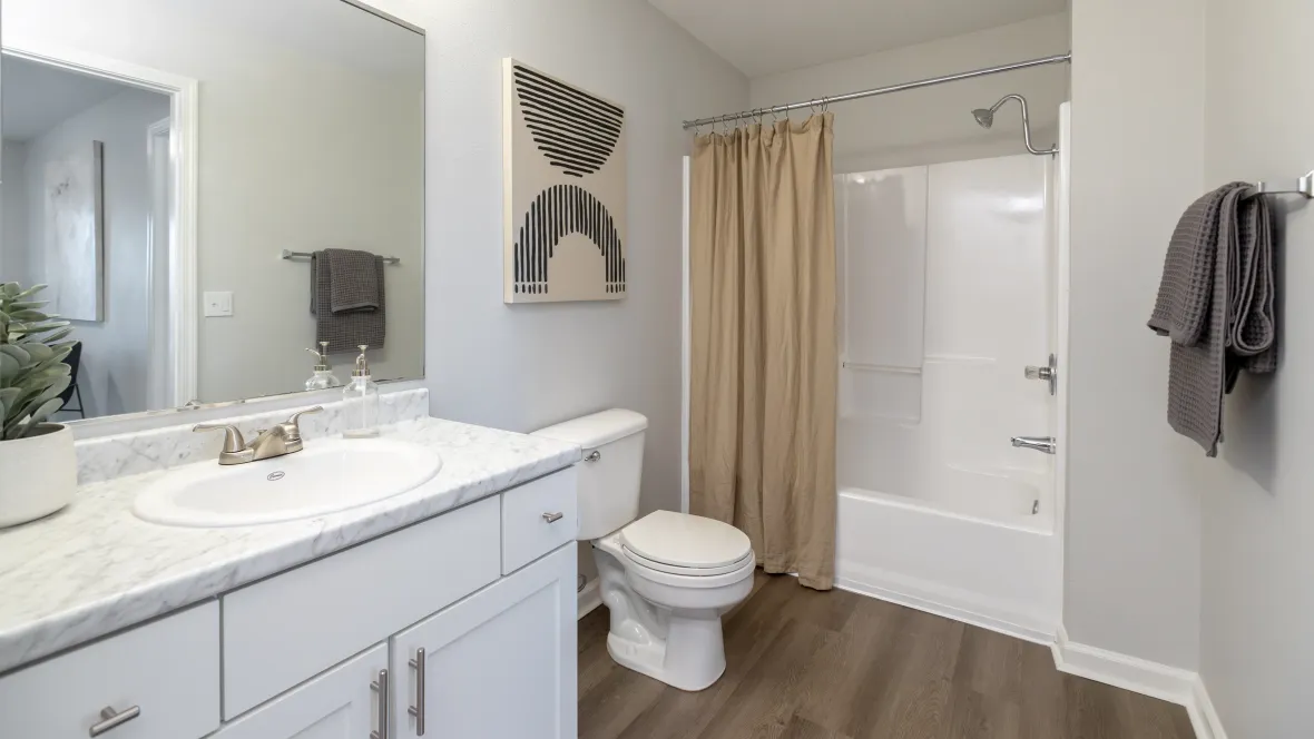  A master ensuite bathroom showcasing an oversized mirror along with a spacious white Carrara countertop with under counter drawers and cabinets boasting optimal storage as well as appealing wood-like floors leading to the shower/tub combo. 