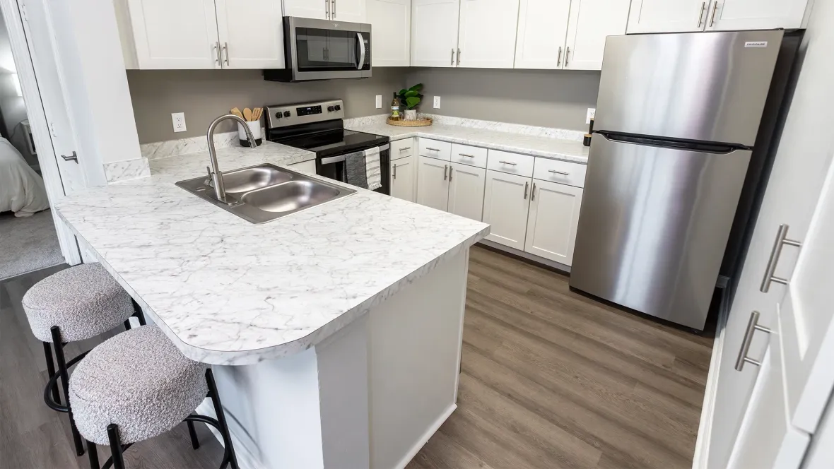 A stylish kitchen space with timeless white marble-style counters accented by a gleaming stainless-steel refrigerator and mounted microwave, and plenty of cabinet storage lining two sides of the kitchen.