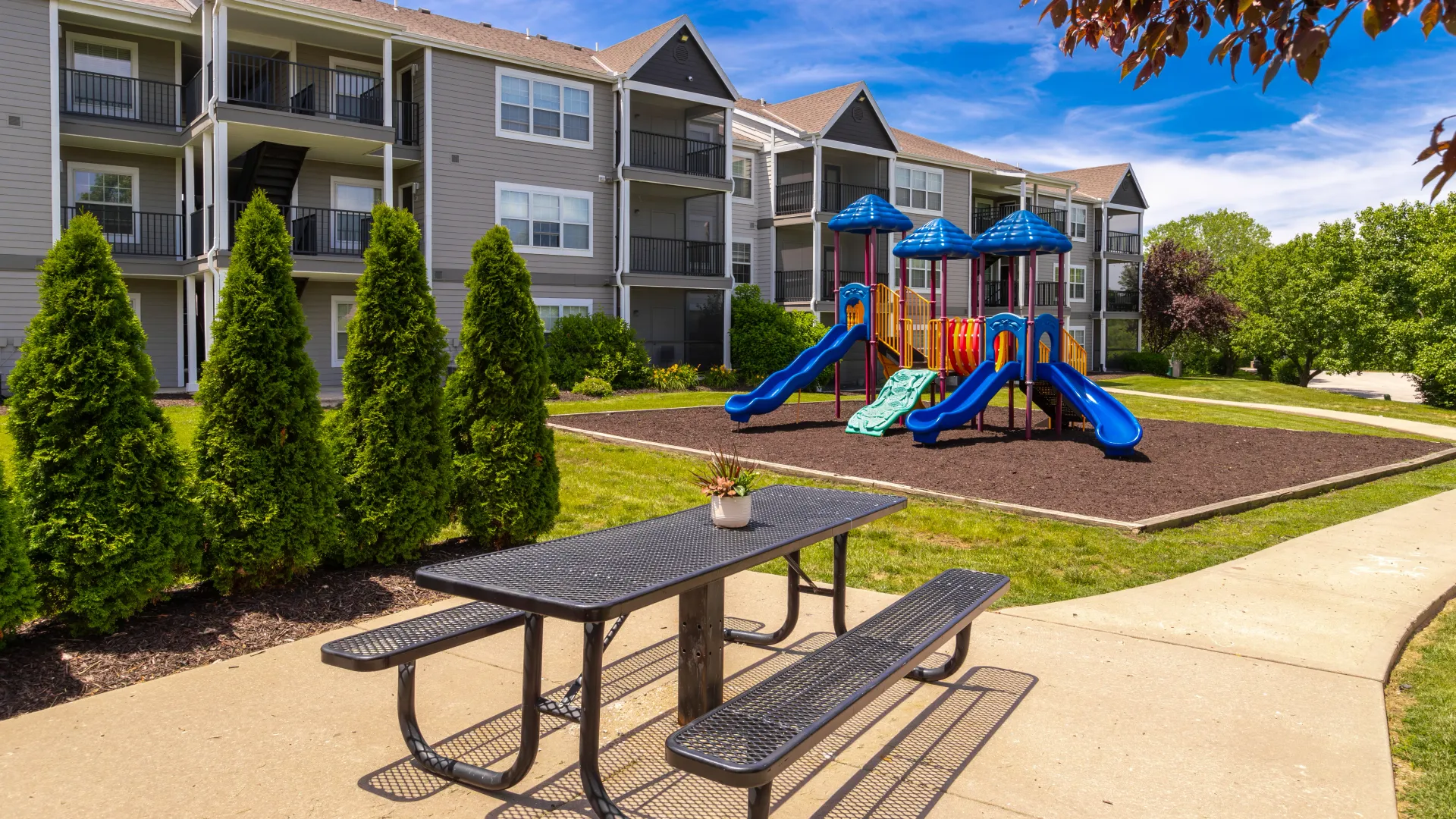 Outdoor playground with blue slides and a picnic table in front of apartment buildings.