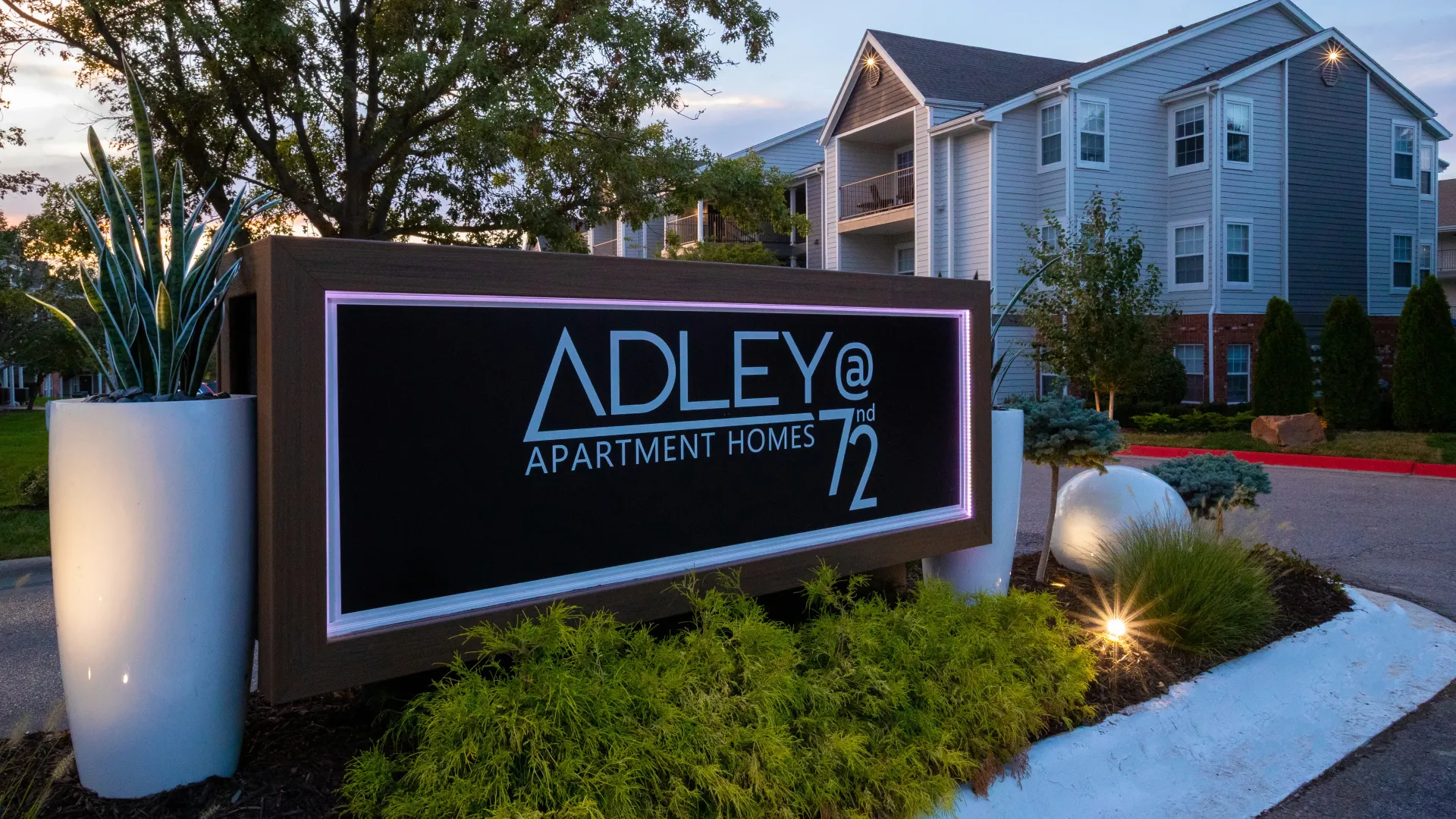 The front sign at the community entrance that reads: "Adley at 72nd Apartment Homes" at dusk, lit with accent lighting.