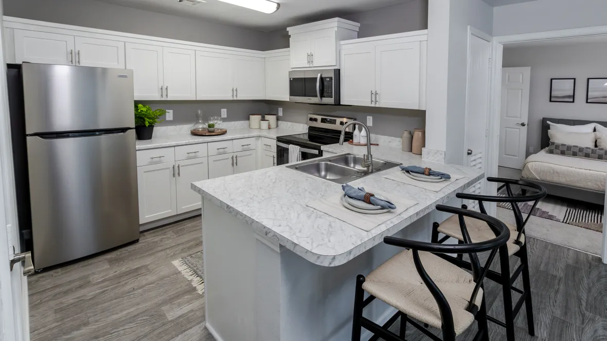 A well-lit kitchen with pristine white cabinetry, white Carrera countertops, a breakfast bar offering overhang for two barstools, wood-style flooring, and stainless-steel appliances.