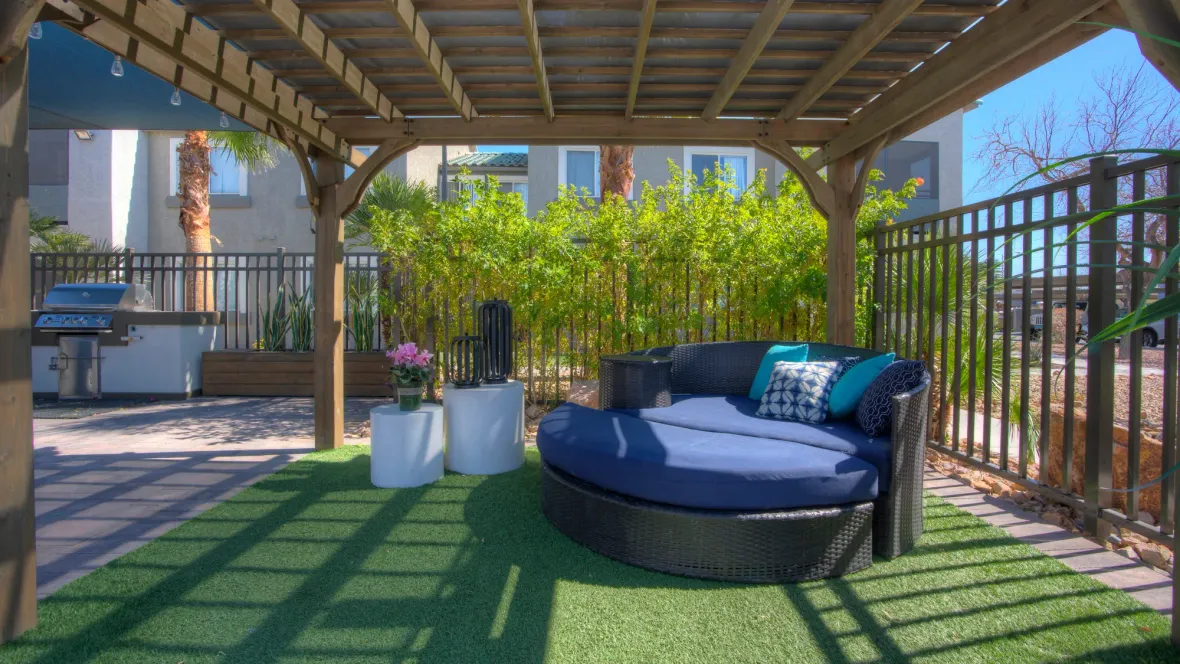A large round, plush lounger beneath a poolside pergola with faux grass carpet underfoot with a backdrop of lush green shrubbery opposite the pool gate, staging a luxurious retreat.