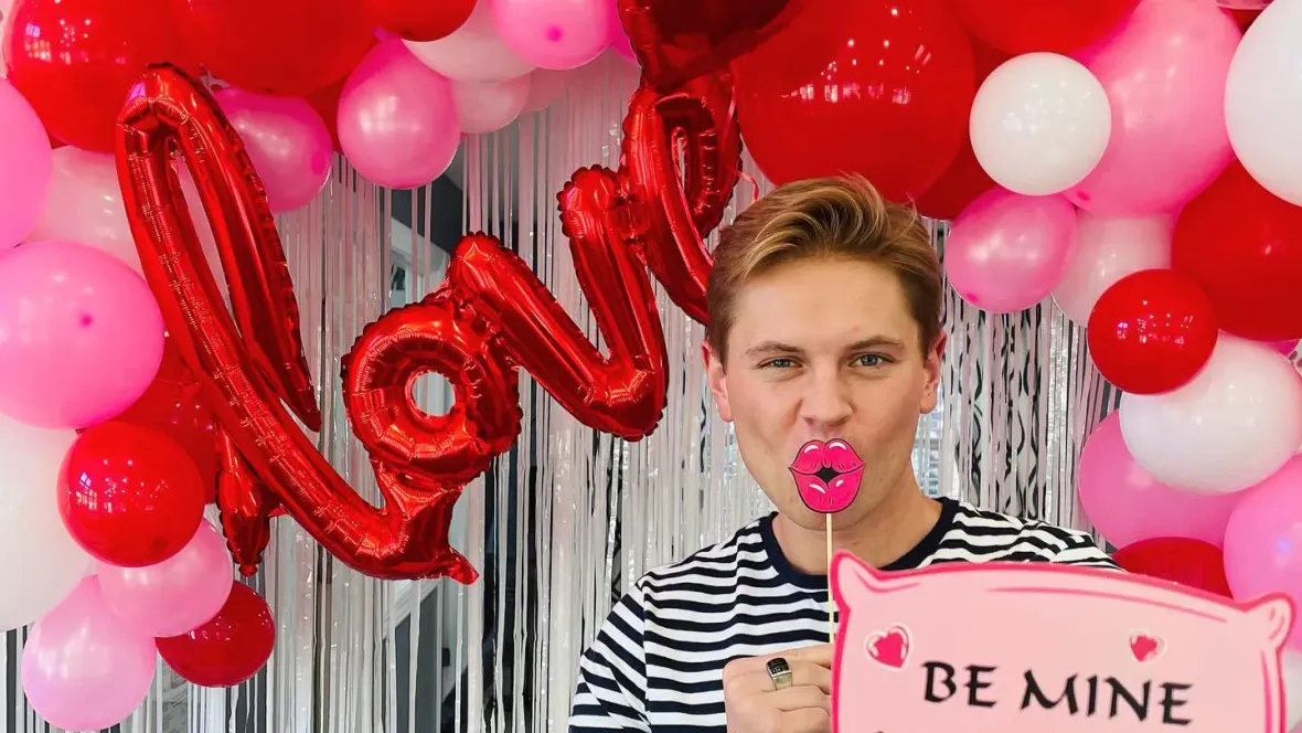 A man in a striped shirt at our Valentine's Day photobooth surrounded by a balloon arc made up of red, pink, and white balloons with a balloon that reads "love" behind him.