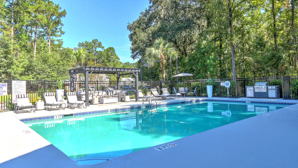 Sparkling swimming pool with a gas grill to the right and multiple loungers lined up along the side of the pool as well as a poolside pergola.