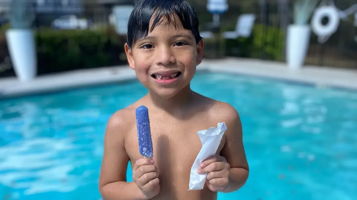 A boy in front of the pool holding a popsicle and smiling.