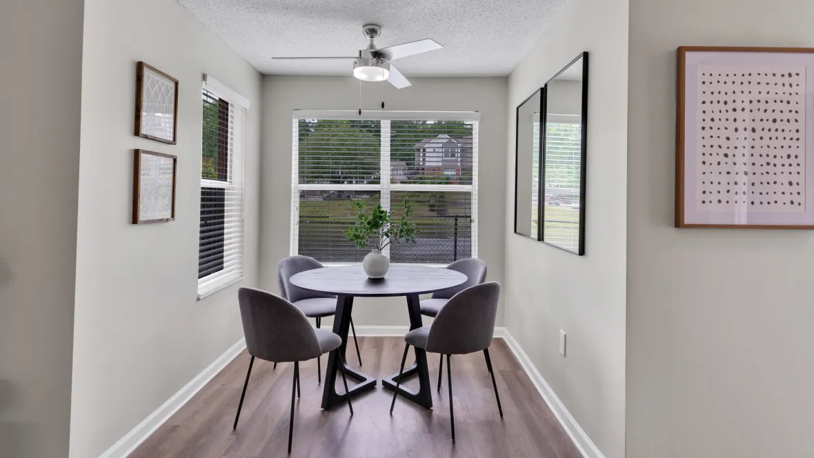 A close-up look at the chic dining space with abundant natural light pouring in through two enormous windows, a dining table, chairs, and an overhead fan.