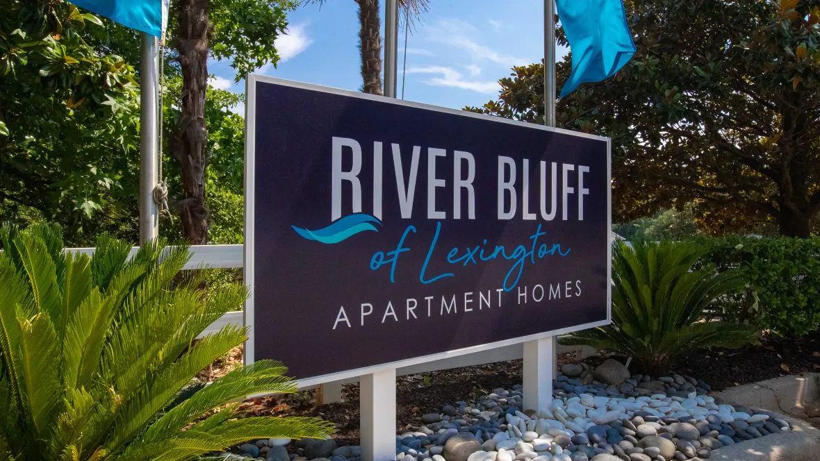 A vibrant welcome guided by the 'River Bluff of Lexington Apartment Homes' entry monument sign adorned with river rocks, lush foliage, and waving blue flags.